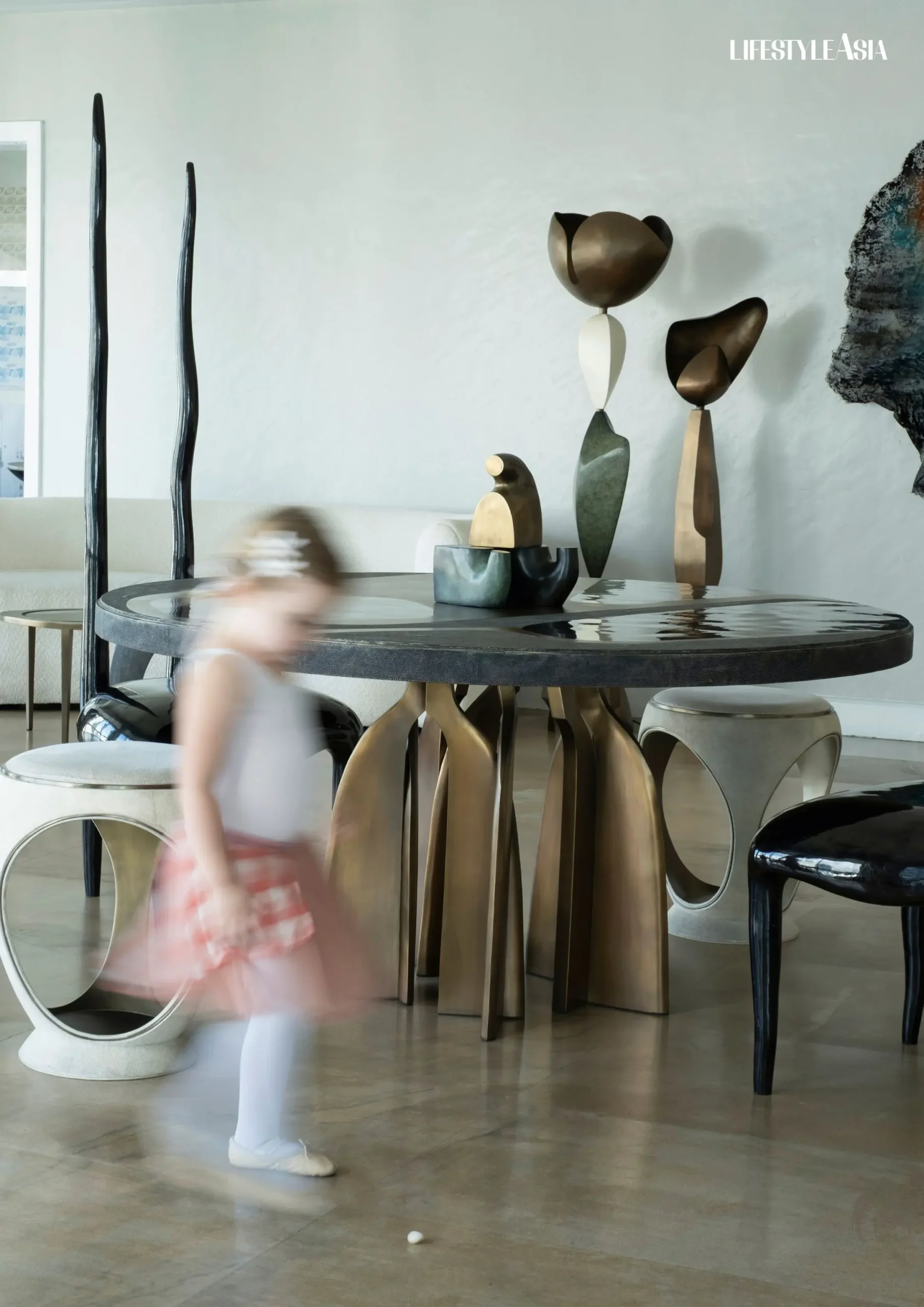 Kifu Augosti and Patrick Coard's daughter River moves through their curated space, where sculptural furniture pieces—including the circular shagreen table with its distinctive base and contrasting round white stools—blur the line between functional design and artistic expression
