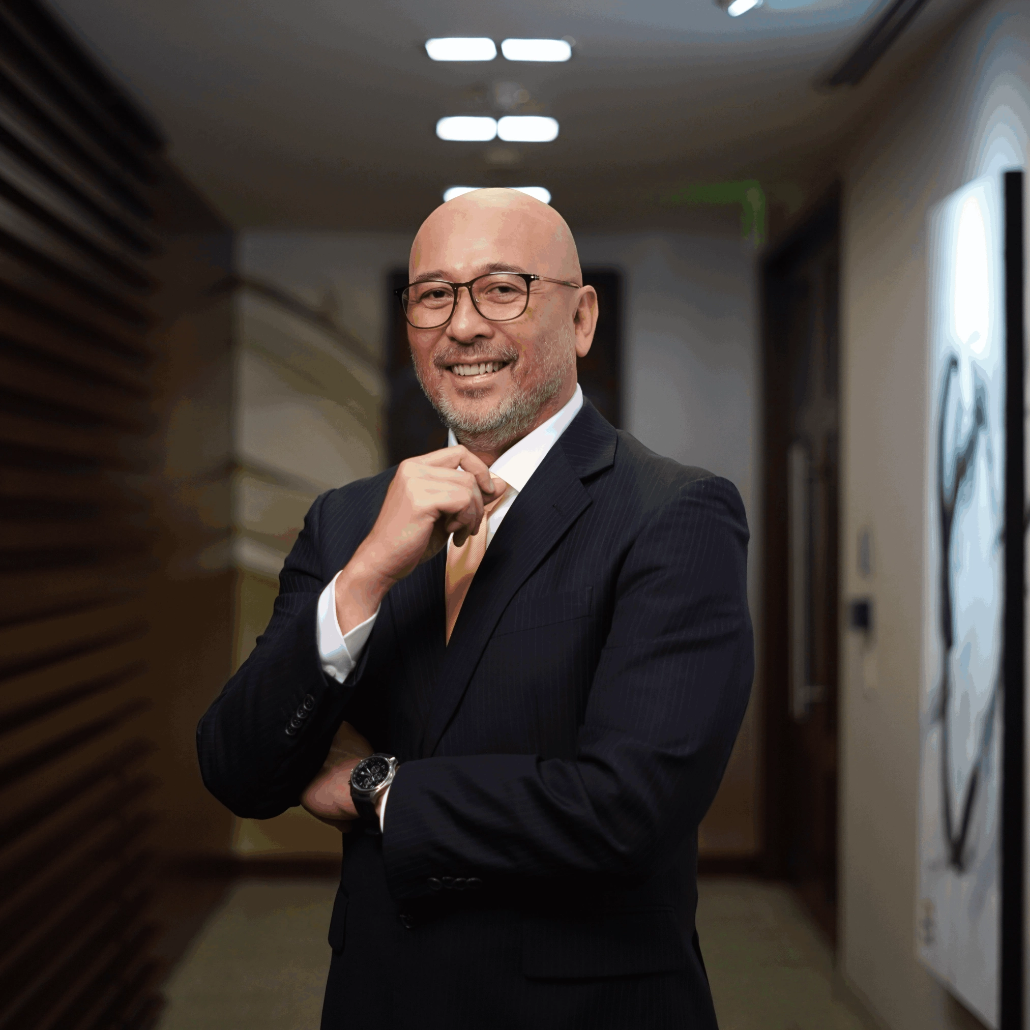Smiling bald man in a dark pinstripe suit, adjusting his tie in a hallway.