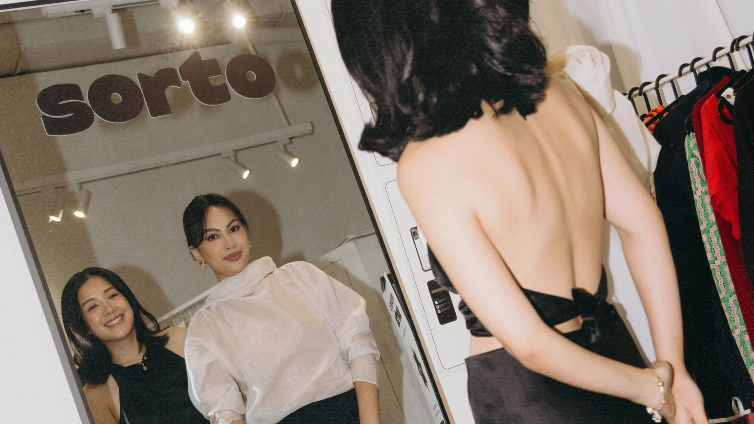 Three women smile while trying on outfits in a clothing store, reflected in a large mirror with racks of colorful dresses nearby and track lights above.