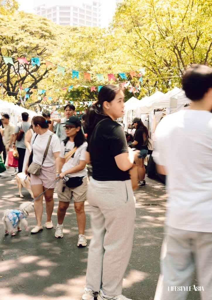 People strolling through an outdoor market with white tents, colorful bunting, and trees on a sunny day.