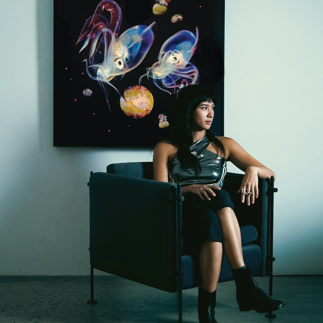 Woman seated in a metallic sleeveless top on a blue chair, with a vibrant jellyfish artwork on the wall behind her.
