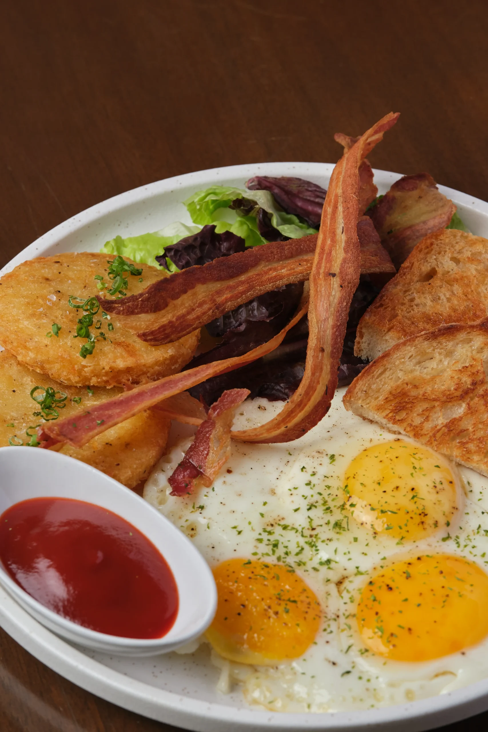 Breakfast plate with three sunny-side eggs, bacon, toast, hash browns, greens, and a side of ketchup.