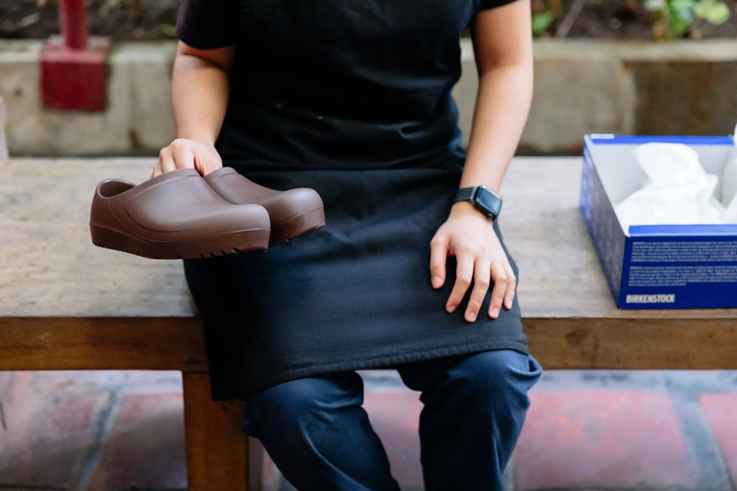 Person wearing a black shirt sits on a wooden bench, holding brown slip-on clogs in one hand with a tissue box nearby.