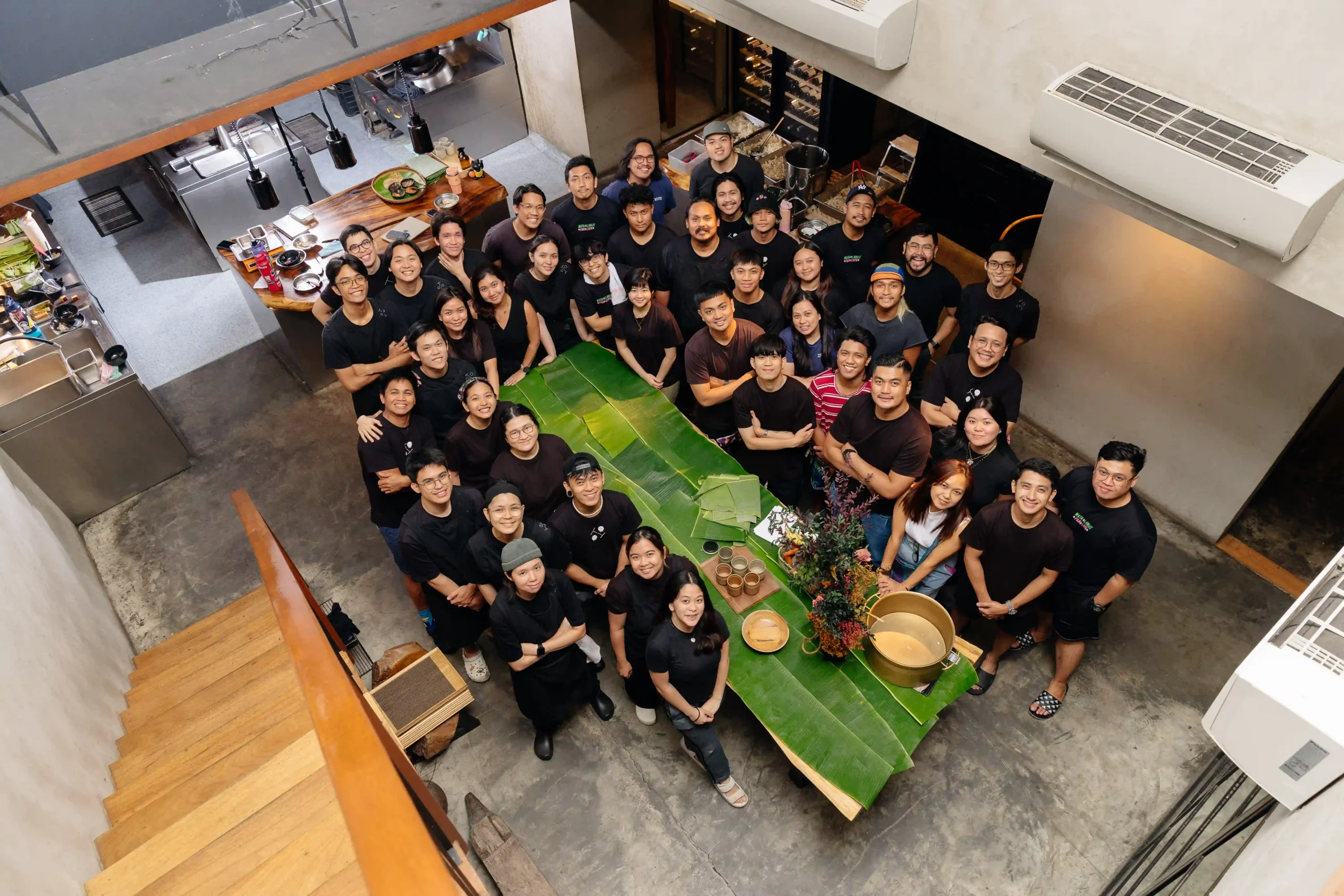 Large group of restaurant staff posing for a team photo around a banana-leaf centerpiece in a modern kitchen/open workspace.