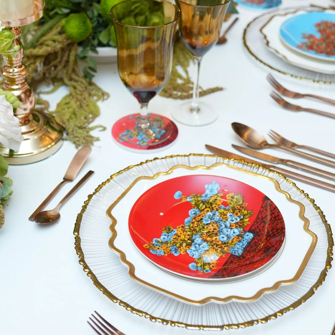 Elegant table setting with a red floral plate atop a white plate with gold trim and a scalloped charger, on a white table with copper utensils and amber goblets.