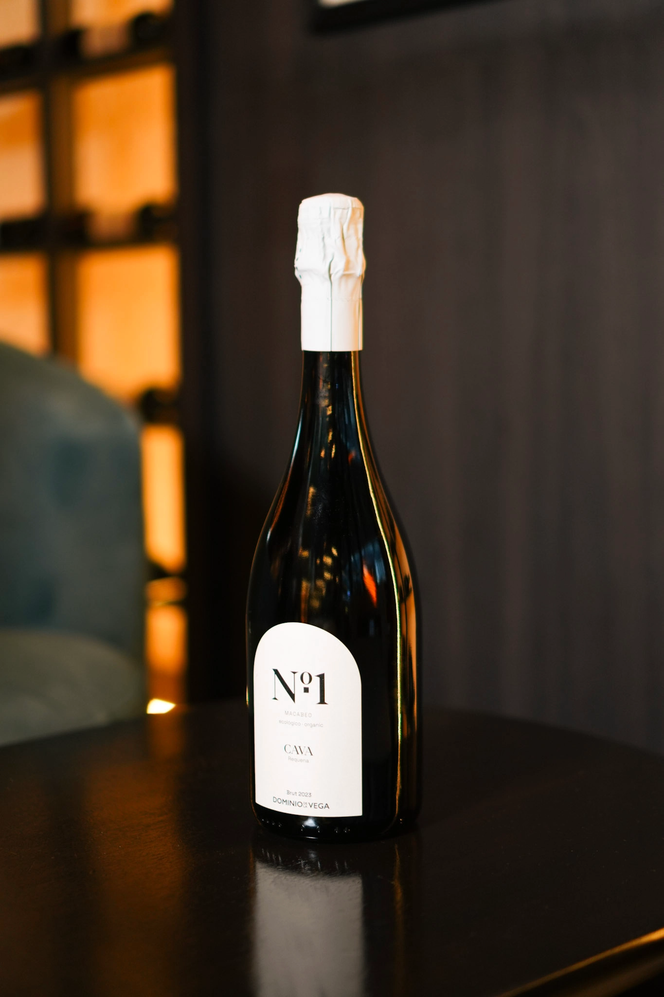 Bottle of No.1 Cava on a dark table with white foil, warm amber background, label showing 'No.1' and 'CAVA' 2023 Brut.