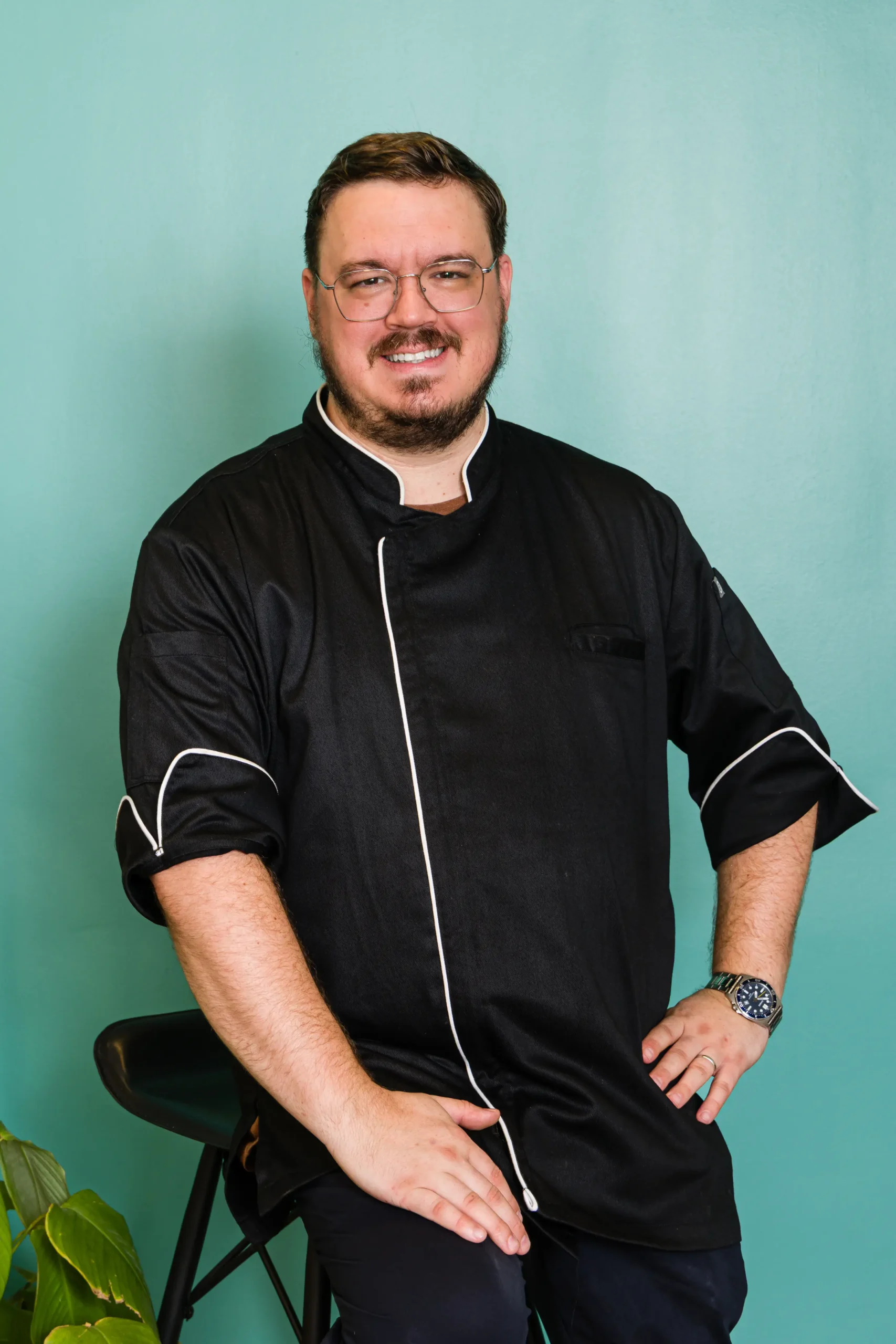 Smiling male chef with glasses wearing a black chef coat, seated against a teal wall with white piping on the uniform edge.