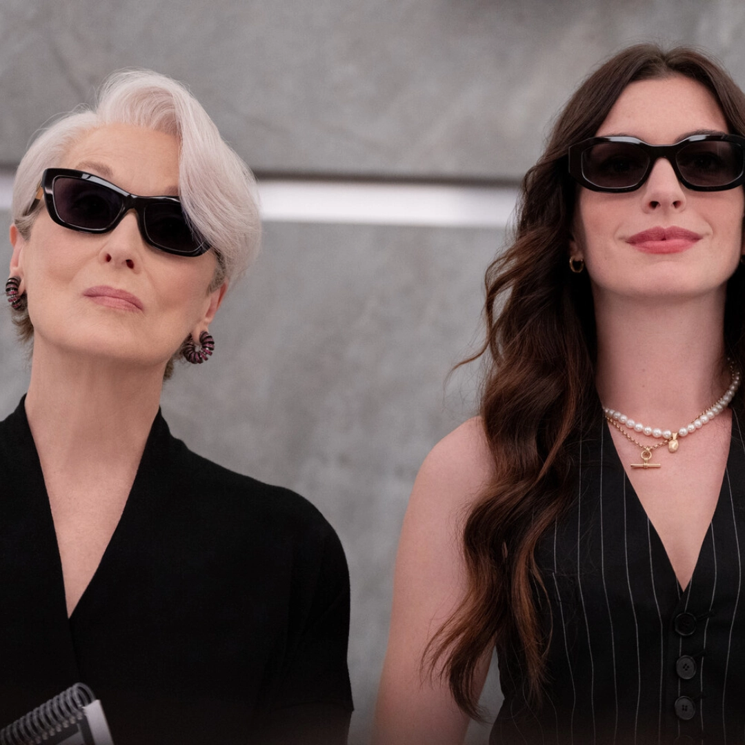 Two women wearing black outfits and sunglasses pose against a gray backdrop for a fashion shot.