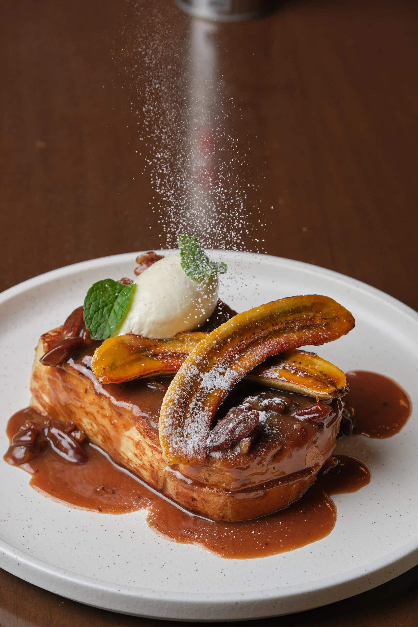 Dessert plate featuring caramel-topped French toast, banana slices, a scoop of vanilla ice cream, mint leaf, and dusted with powdered sugar.