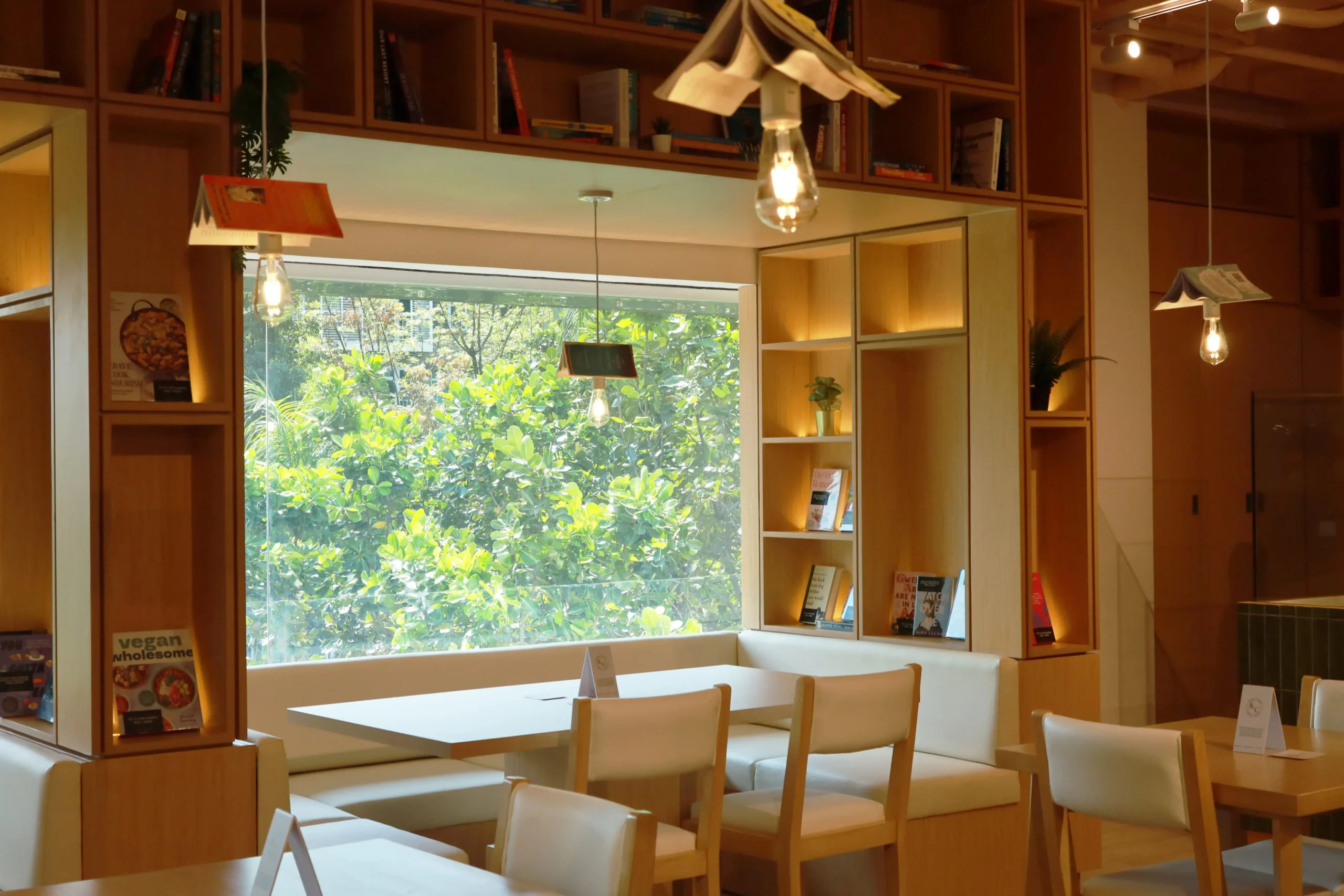 Cozy cafe interior with wooden shelves, warm Edison bulbs, and white tables facing a large window with green plants outside.