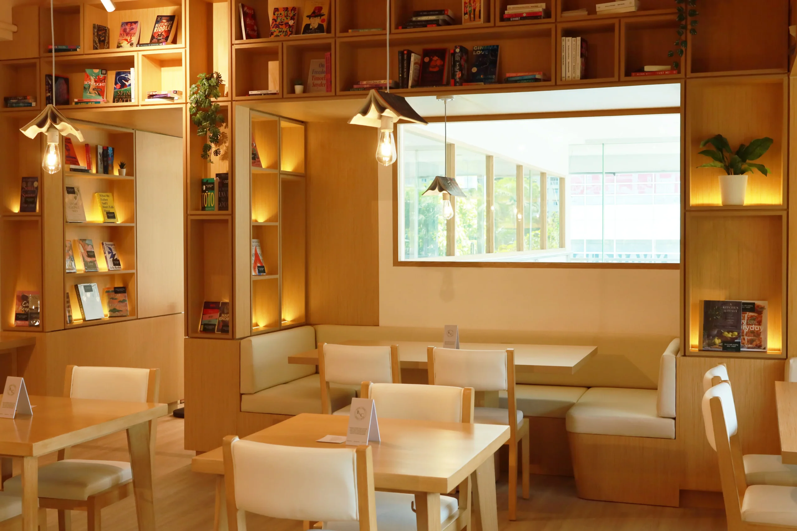Wooden cafe area inside a bookstore with built-in shelves of books along the walls, warm pendant lights, and cream upholstered chairs at light wood tables.