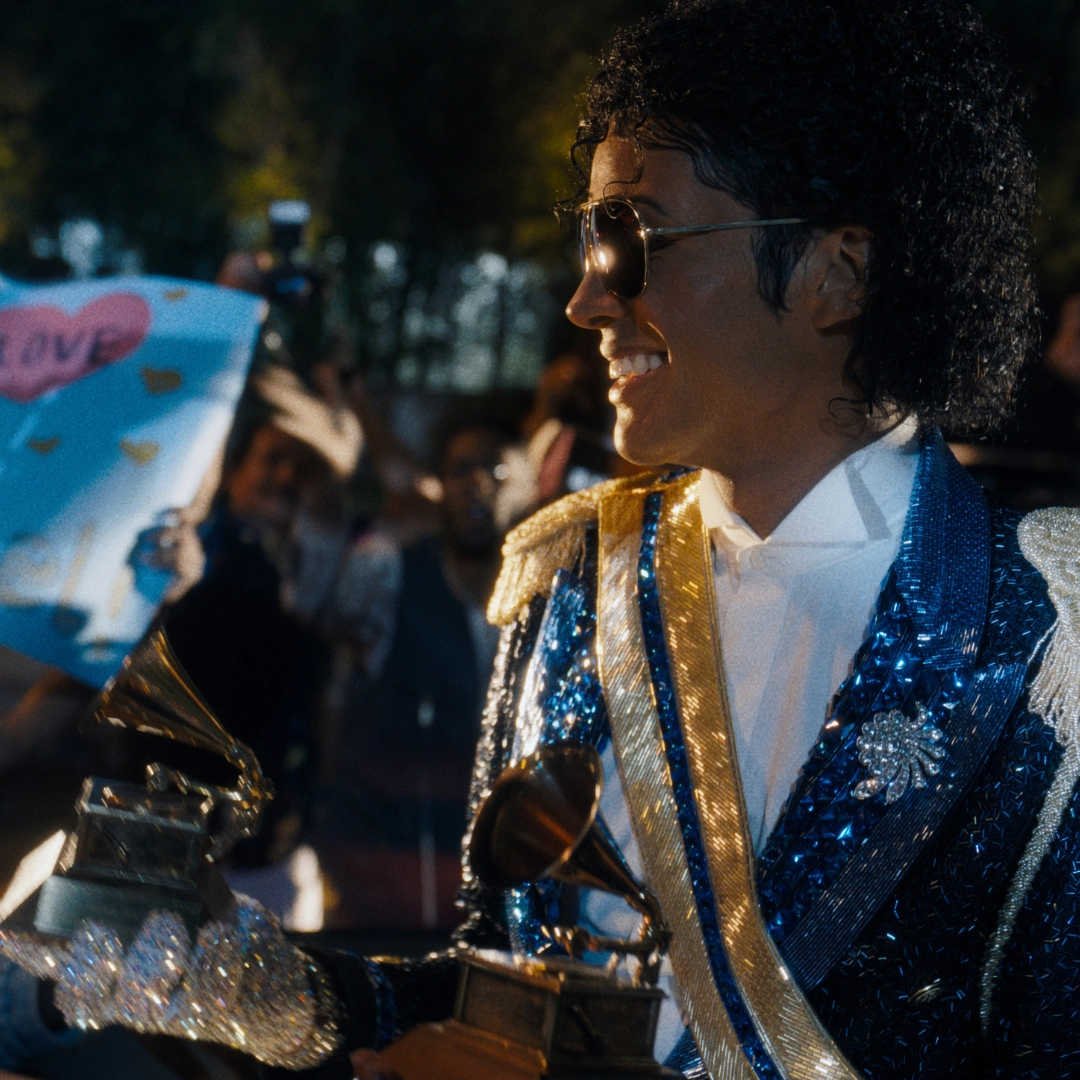 Performer in a sequined blue and gold jacket, wearing sunglasses, smiling while holding a shiny trophy-like award at a nighttime outdoor event. Behind him, a cheering crowd.