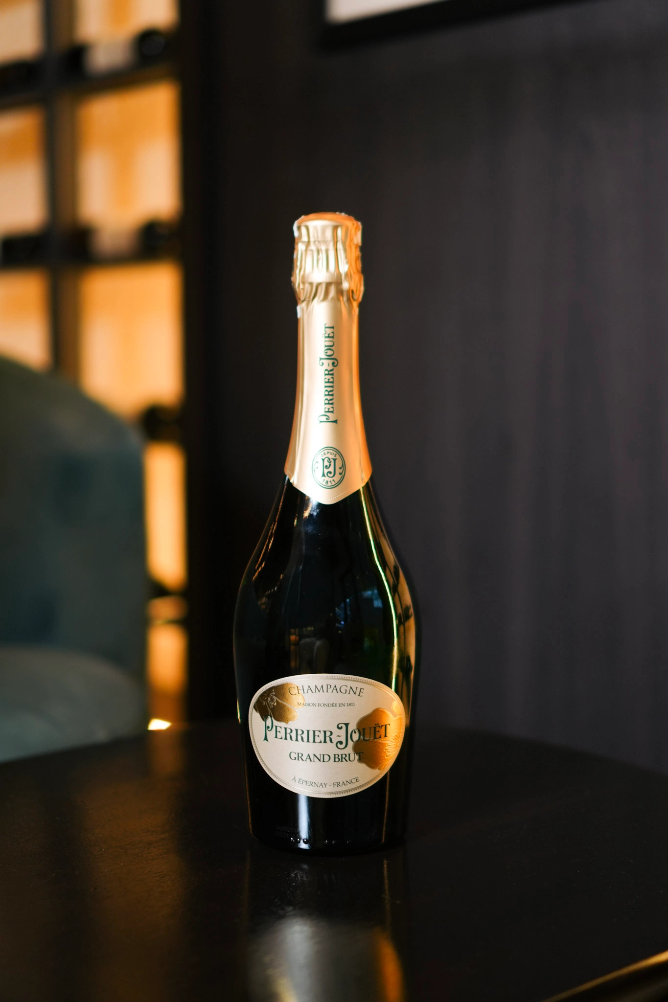 Champagne bottle with gold foil labeled Perrier-Jouët Grand Brut on a dark table in warm lighting.
