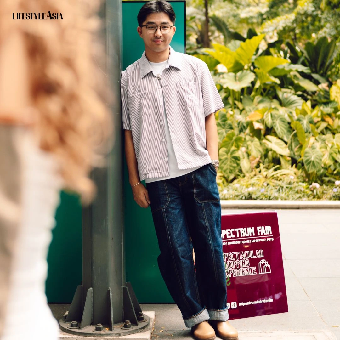 Casual portrait of a man in a striped shirt and rolled jeans leaning against a green pole at an outdoor fair, with tropical plants in the background and a Spectrum Fair sign nearby.