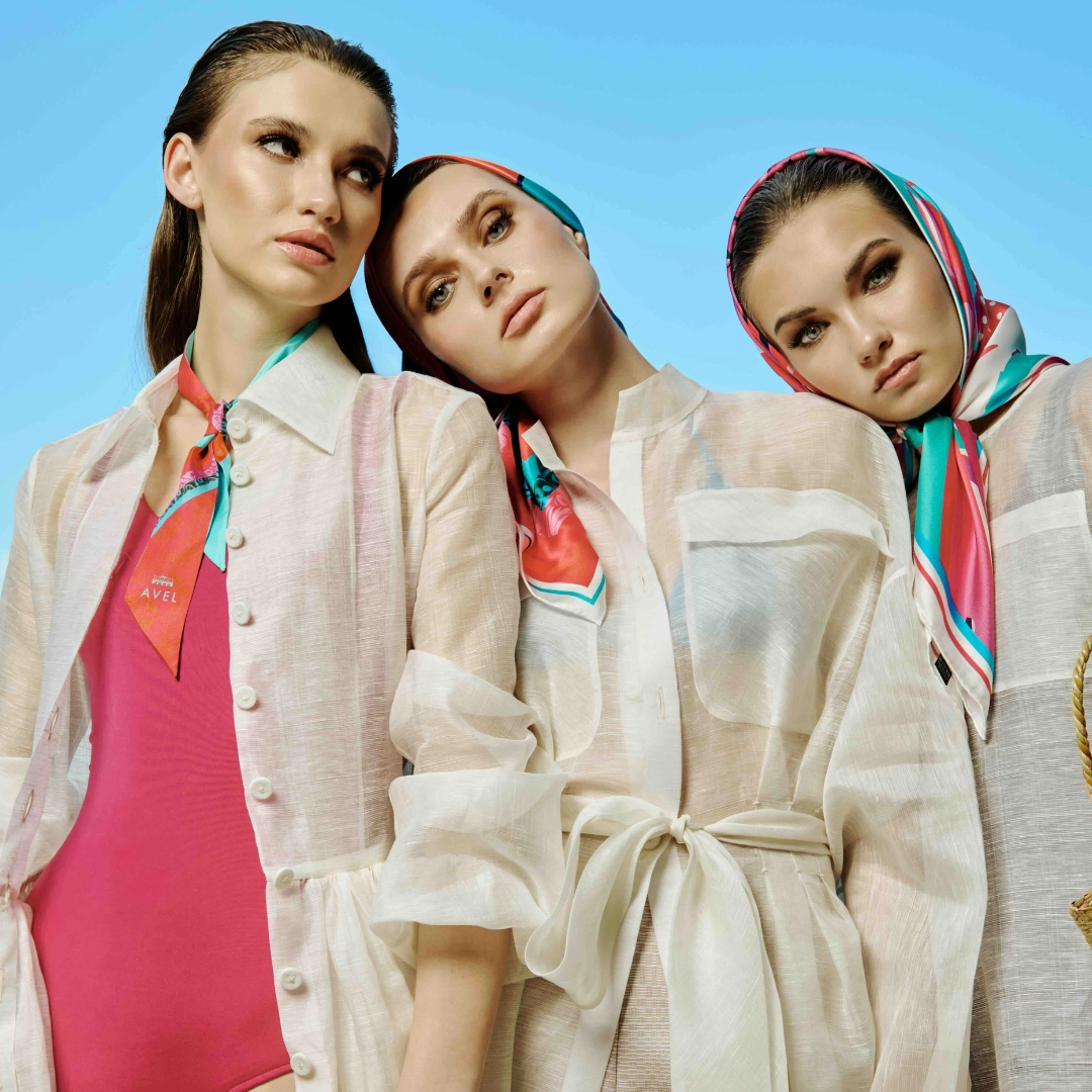 fashion models pose outdoors in front of blue city skyscrapers; woman wears a colorful patterned headscarf and sheer dress