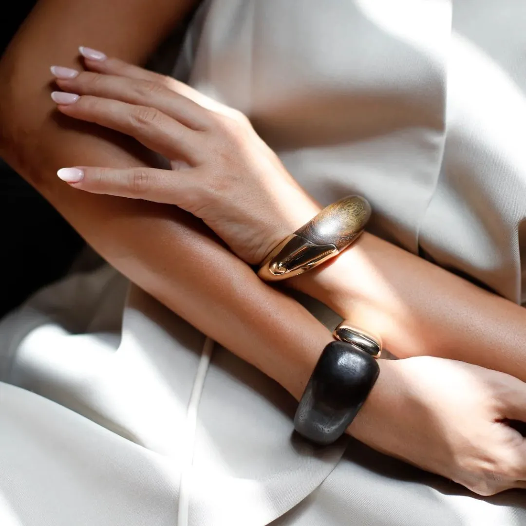 Two forearms crossed, showcasing a gold wide cuff bracelet and a chunky black-gray bracelet against a light outfit.