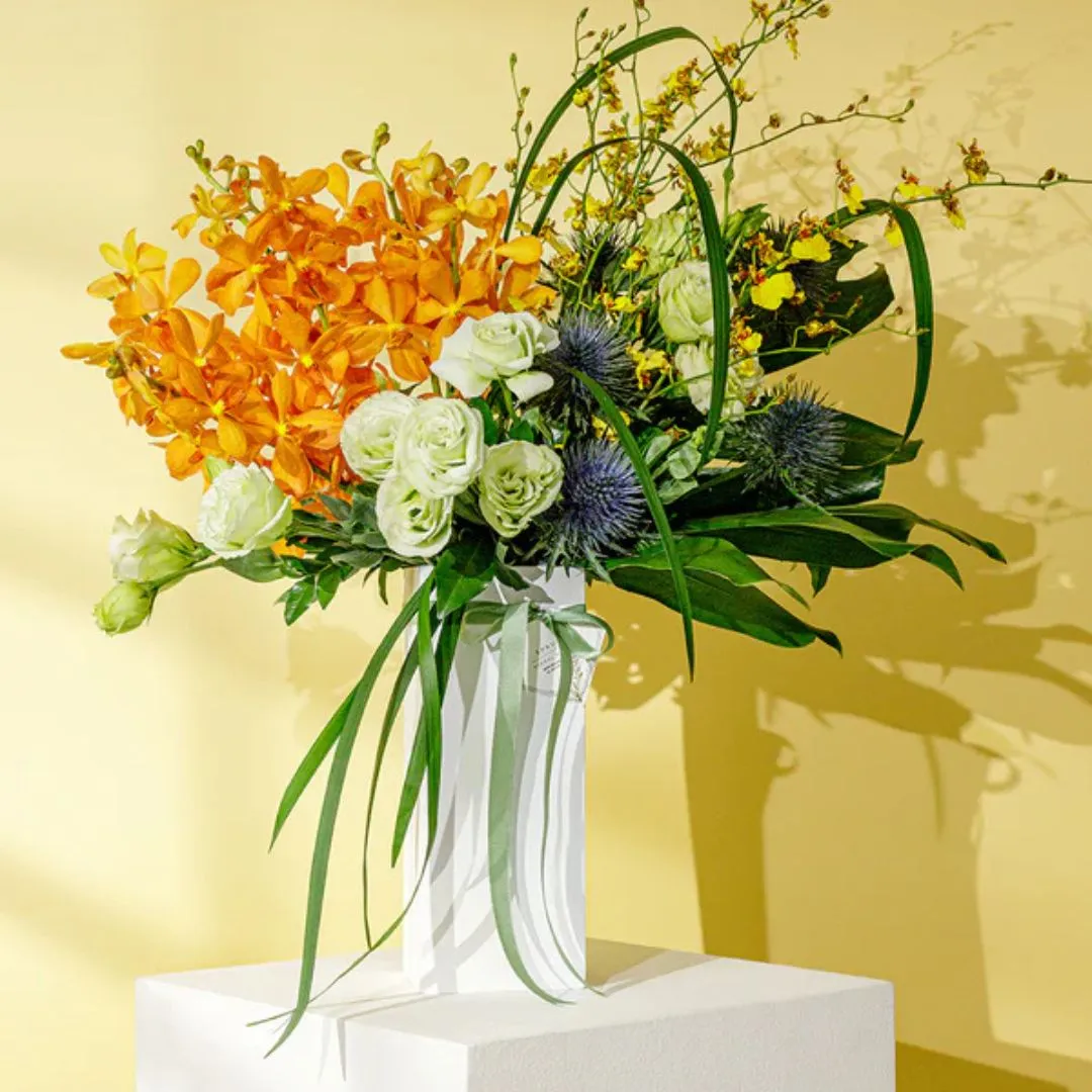 Flower arrangement with orange orchids, white roses and blue thistles in a white vase with green leaves on a pedestal against a pale yellow wall.