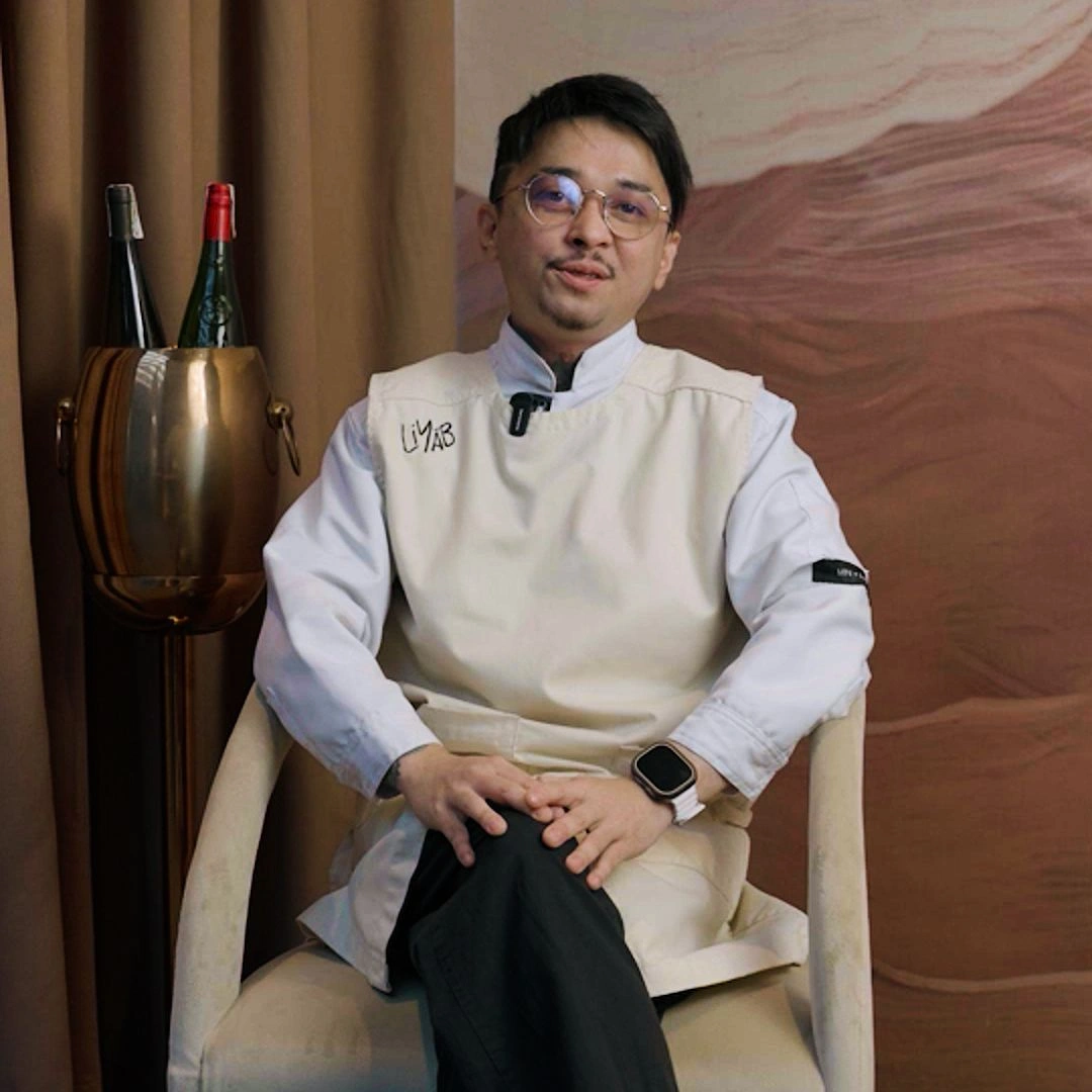 Chef in a white apron sits on a chair with hands folded, wearing glasses and a smartwatch, champagne bottles nearby in a copper bucket.
