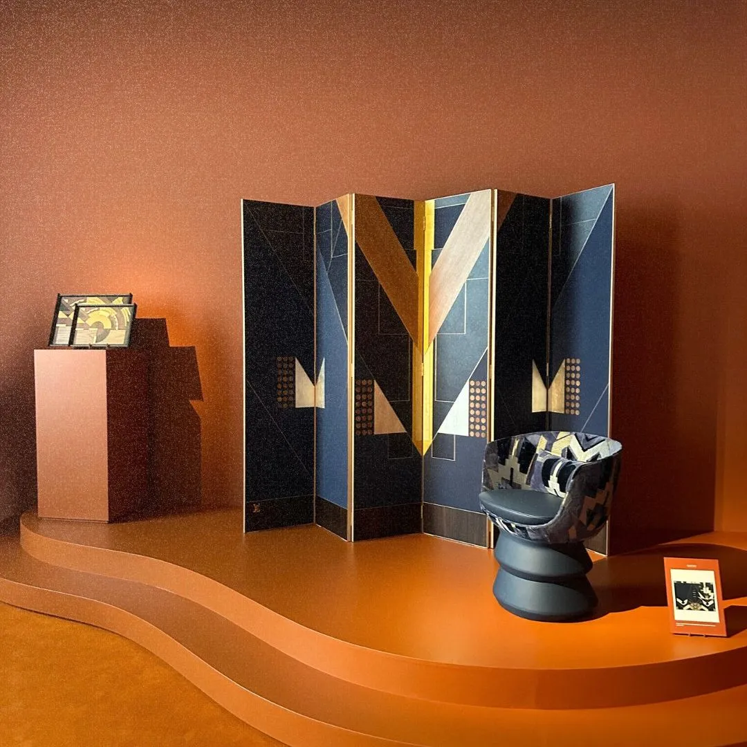 Interior exhibition display featuring a geometric folding screen, a leather-like sculpted chair, and warm orange backdrop on a curved platform.
