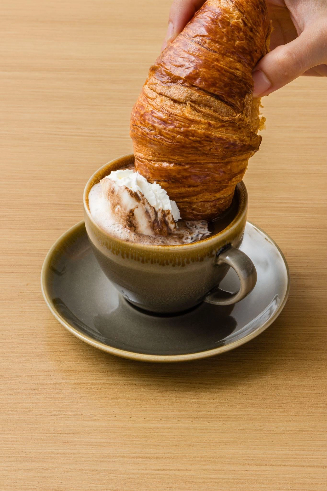 Croissant being dipped into a cup of coffee with whipped cream on a wooden table.