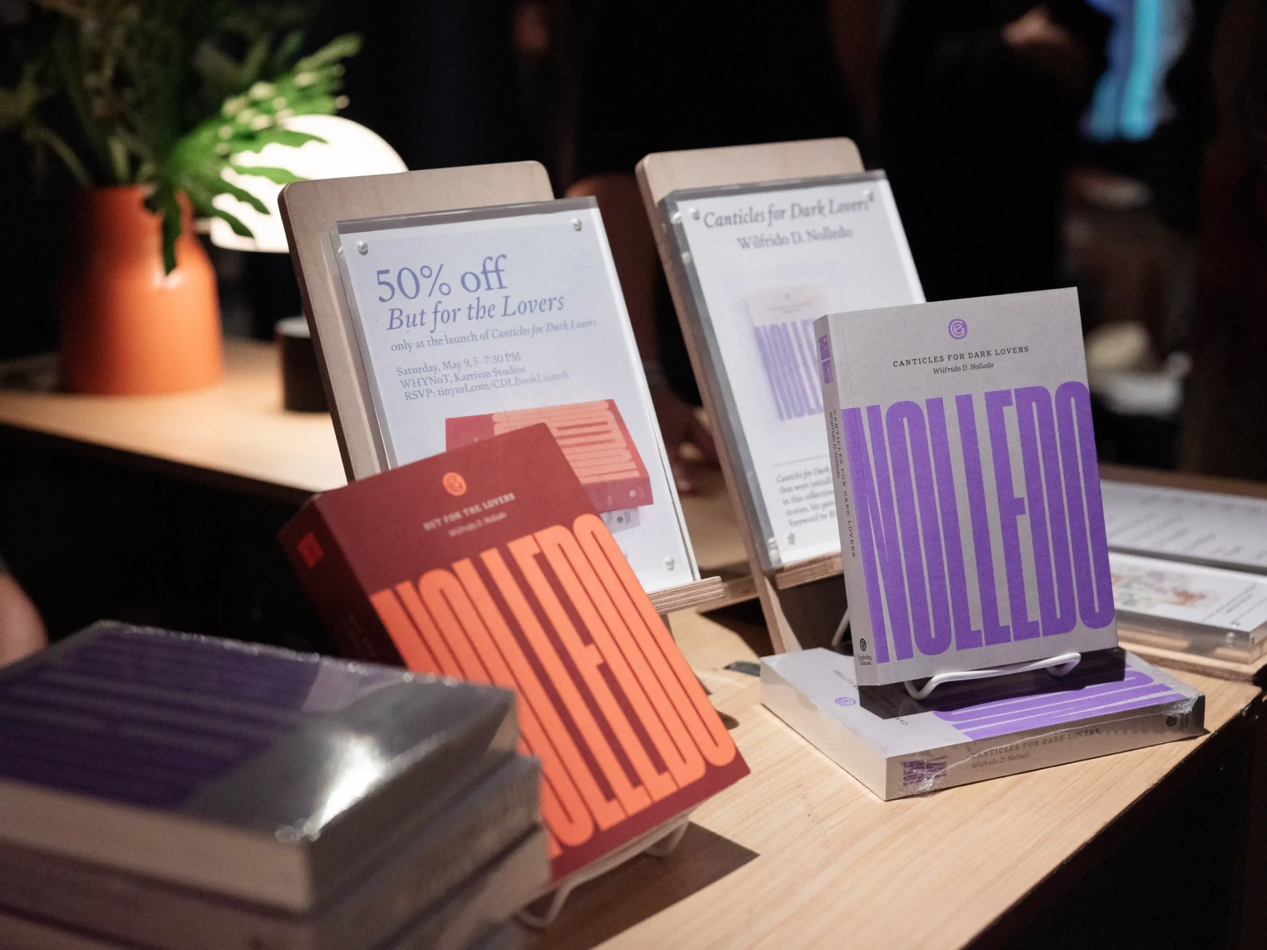 Display of promotional booklets on a wooden table; orange and purple covers with large text, arranged in stands with a plant in the background.