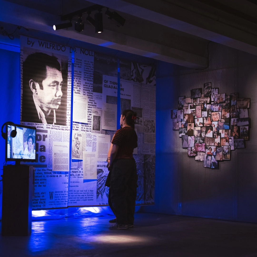 Person standing and viewing a newspaper-wall installation with a large black-and-white portrait, blue-lit gallery.