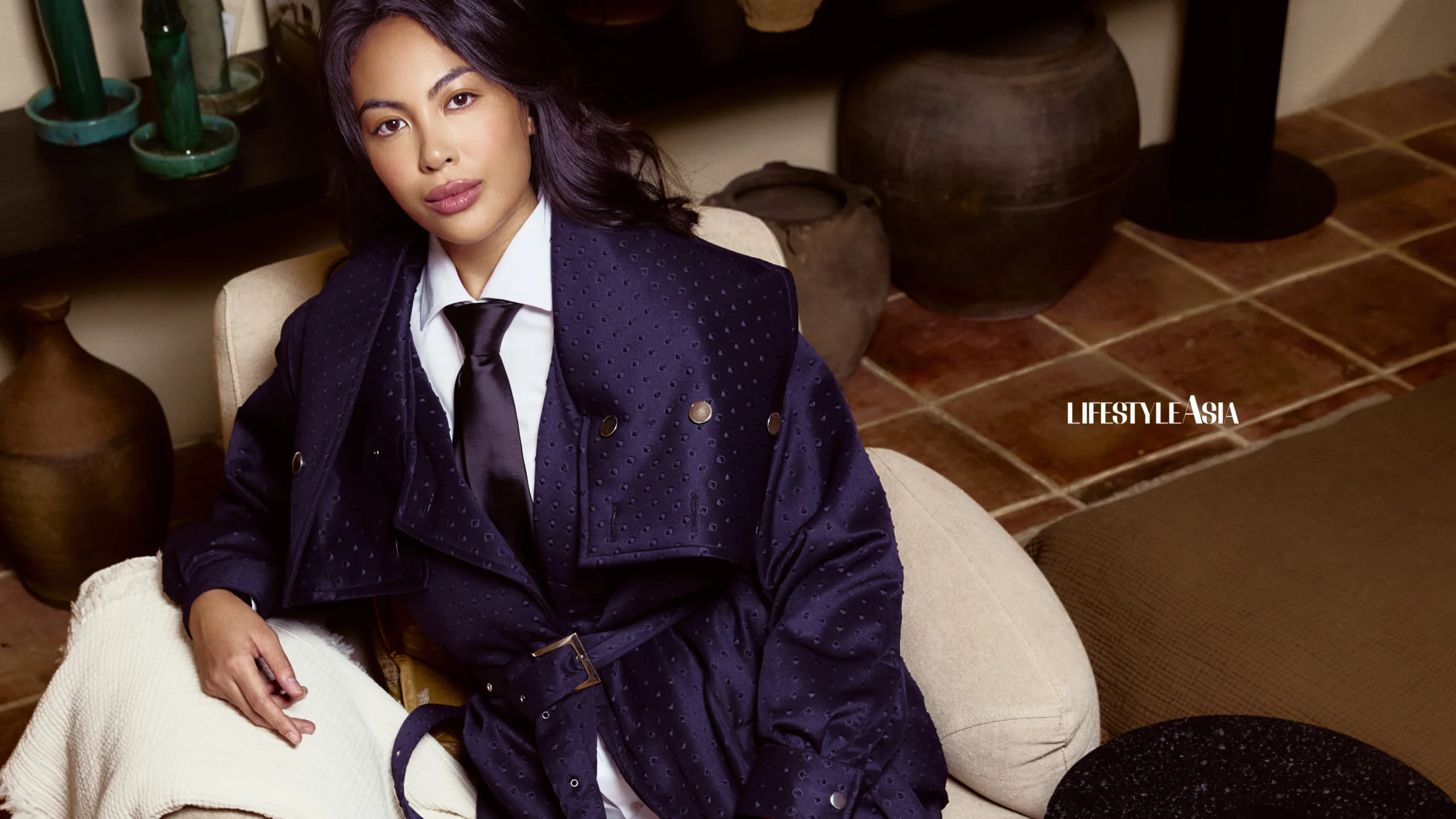 Woman in a navy dotted blazer and white shirt seated on a cream sofa, with pottery and tiled floor in a stylish interior (Lifestyle Asia watermark).