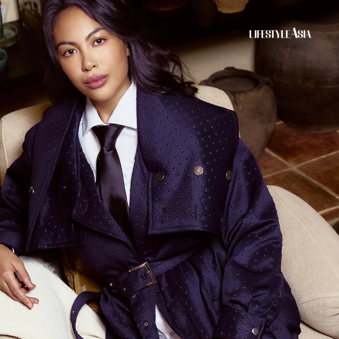 Woman in a navy dotted trench coat with a white shirt and black tie sits on a beige chair for a lifestyle photoshoot; Lifestyle Asia logo in the corner.