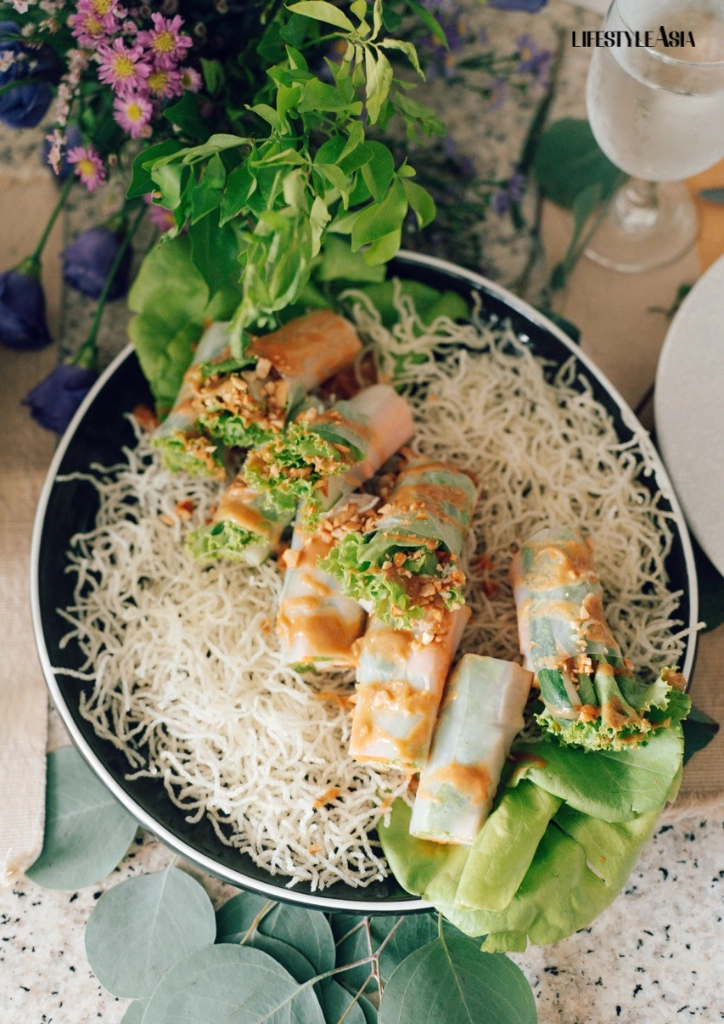 Plate of fresh veggie spring rolls with sesame crunch atop a nest of rice noodles, garnished with herbs on a dark platter.