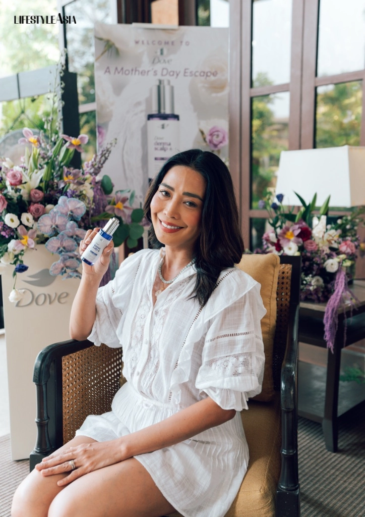 Woman in a white dress sits on a chair indoors, holding a Dove skincare bottle with a promotional banner and floral displays in the background.