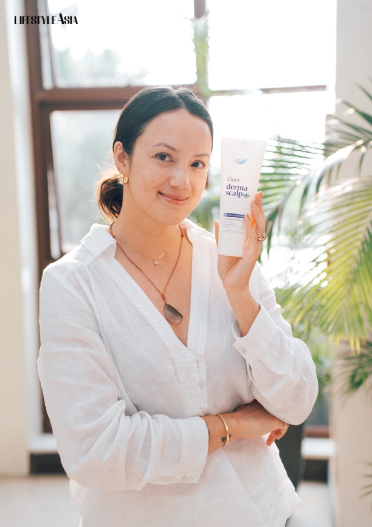 Smiling woman in a white blouse holds a Dove DermaScalp tube in a bright, plant-filled room.