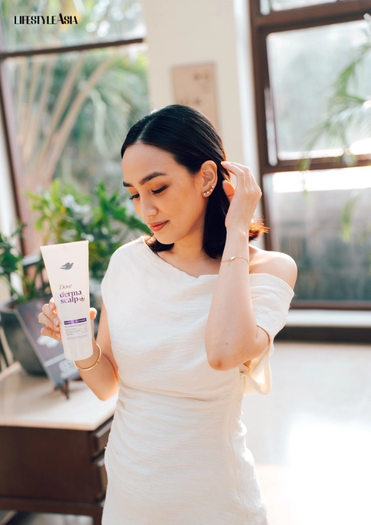 Woman in a white off-shoulder dress holds a Dove DermaScalp shampoo bottle in a bright, plant-filled room; Lifestyle Asia logo visible in the corner.