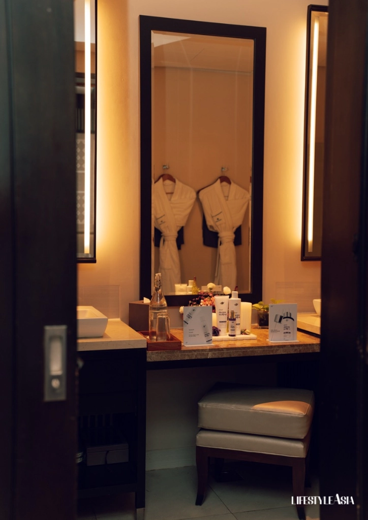 Hotel bathroom vanity with a framed mirror, two white robes on hangers reflected in the mirror, and toiletries on the counter.