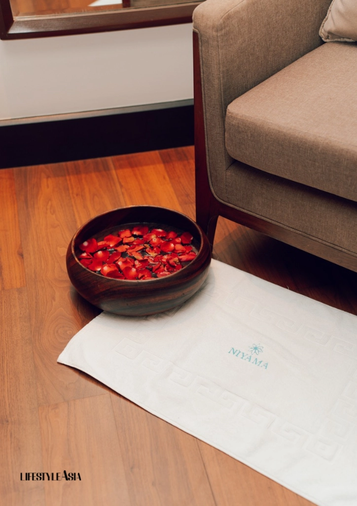 Dark ceramic bowl filled with floating red rose petals rests on a white spa towel beside a beige sofa on a wood floor, suggesting a relaxing spa setting.
