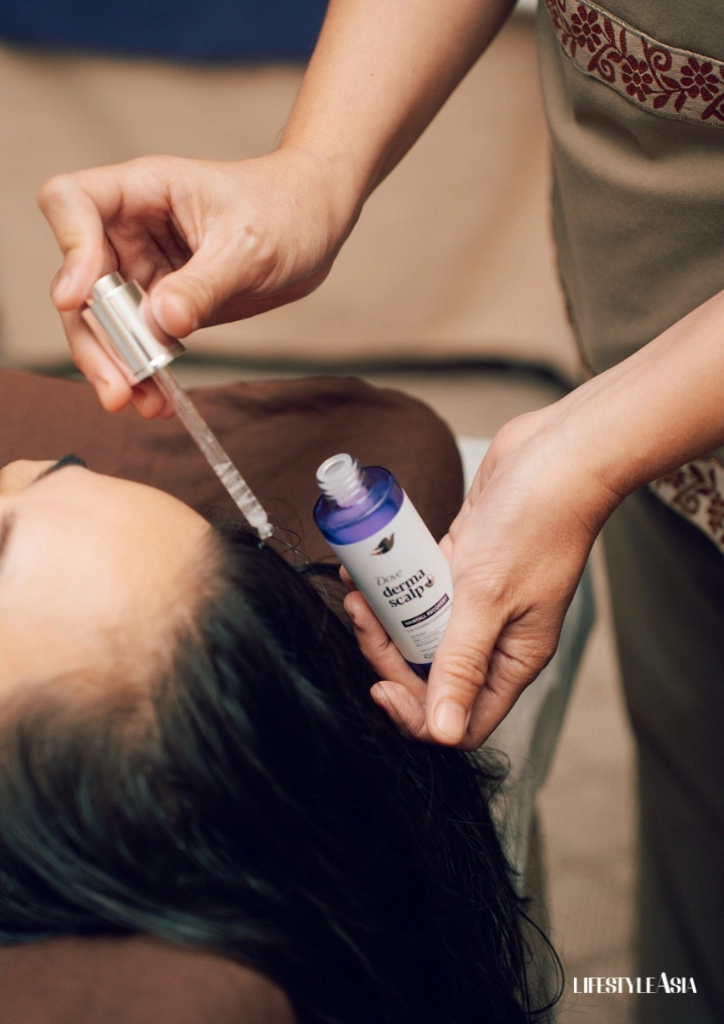 Practitioner using a dropper to apply a liquid to a client's scalp; blue bottle labeled Derma Scalp sits nearby.
