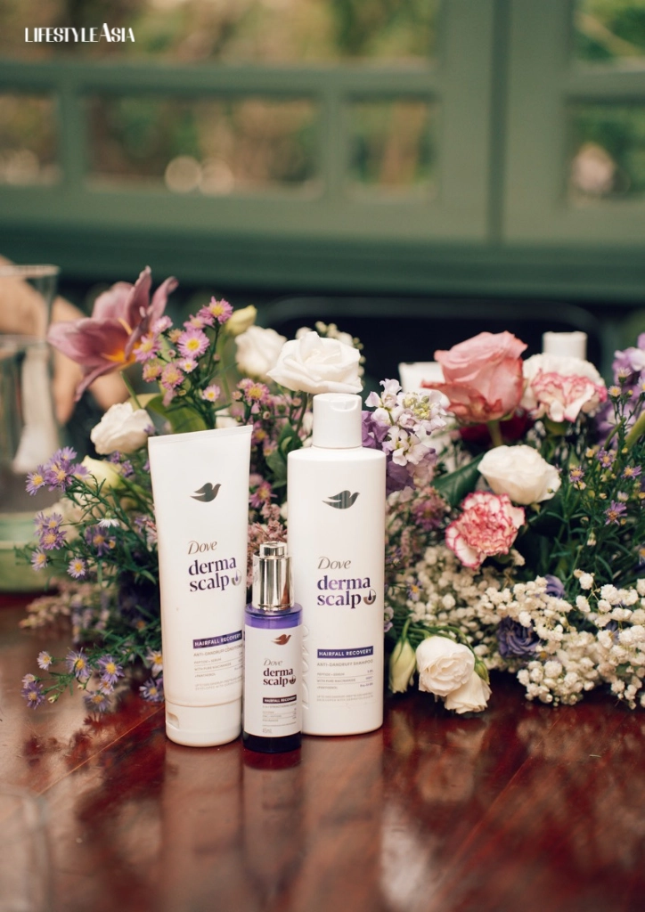 Dove DermaScalp haircare bottles with a purple-topped bottle among pink and white flowers on a wooden table.