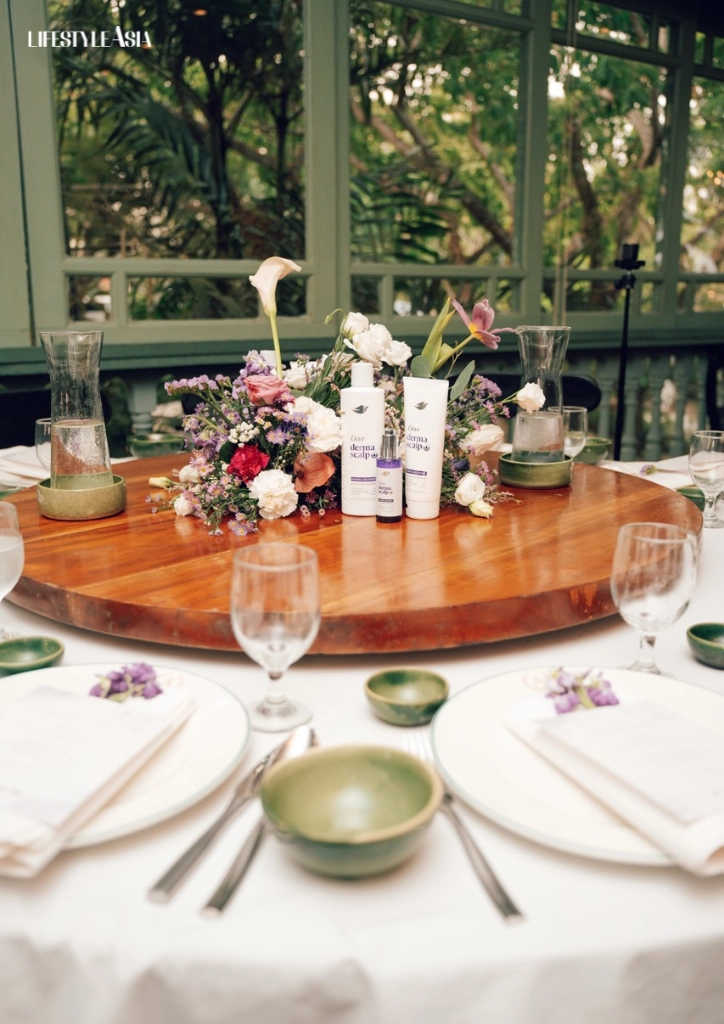Table set for a formal event with a flower centerpiece and skincare product bottles on a round wooden tray at the center of the table.