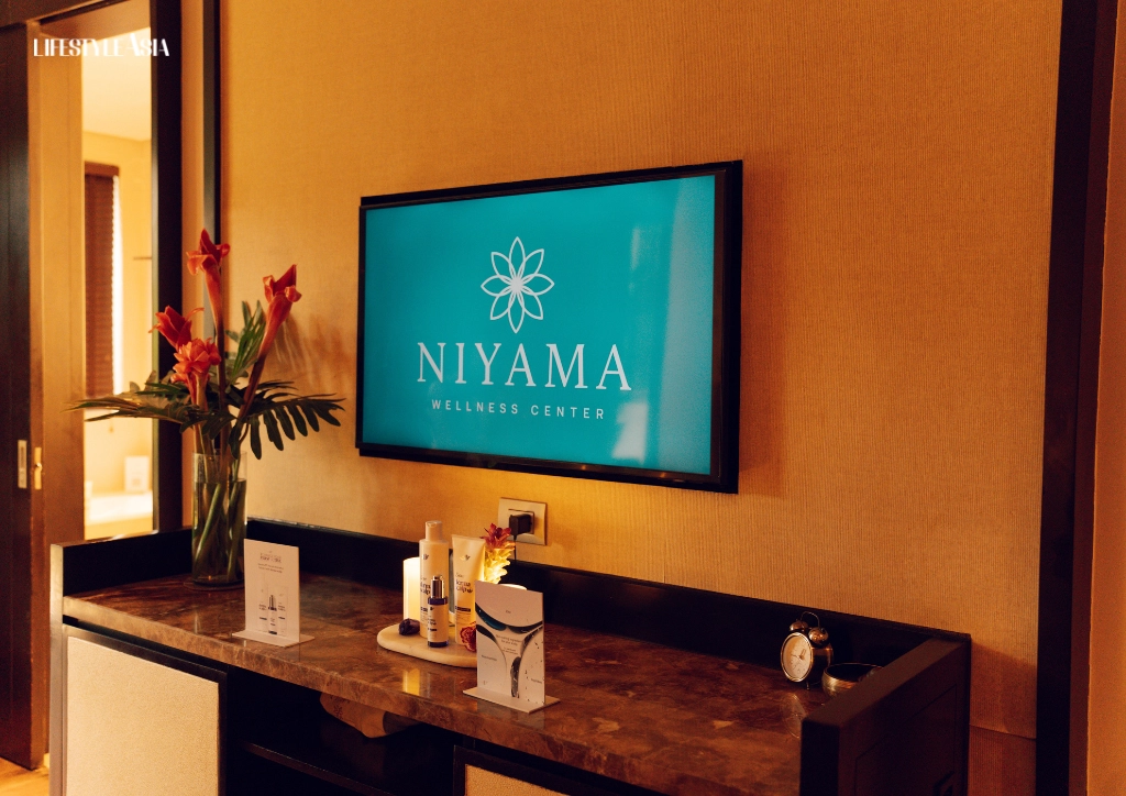 Teal NIYAMA Wellness Center logo on a wall-mounted screen above a marble counter with red flowers and spa products.