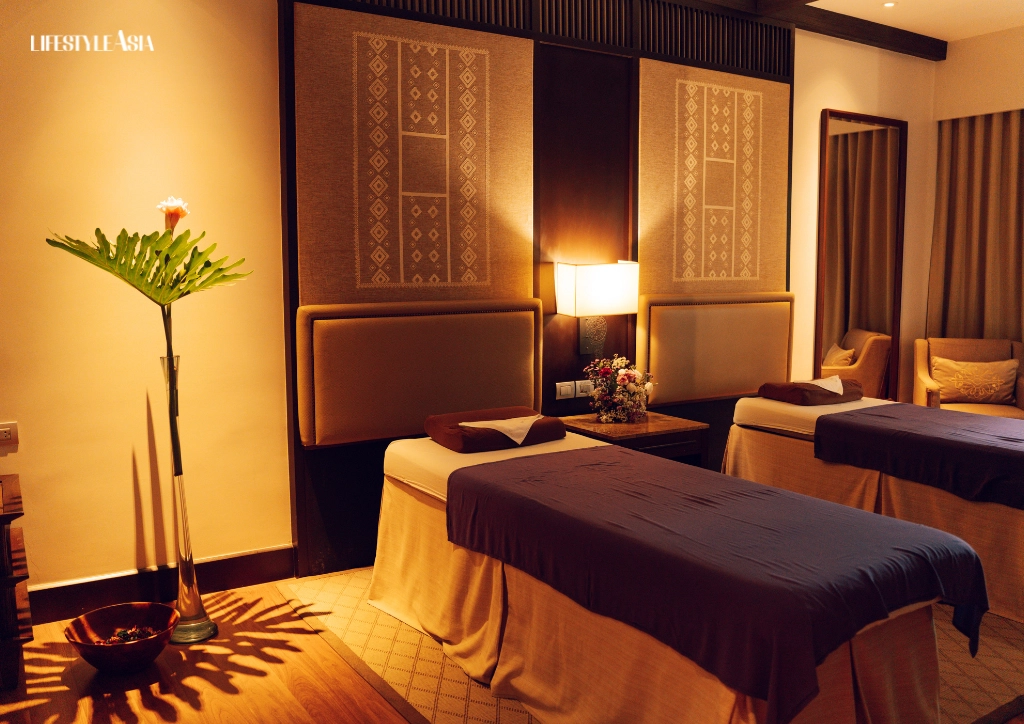 Dimly lit hotel spa room with two massage beds, beige linens, and a tall plant by the left wall.