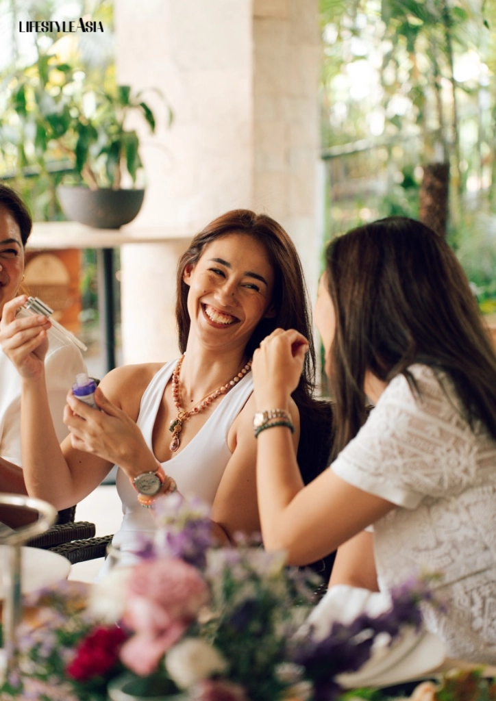 Woman in a white tank top laughs while holding a small bottle at an outdoor brunch among friends in a sunlit setting.