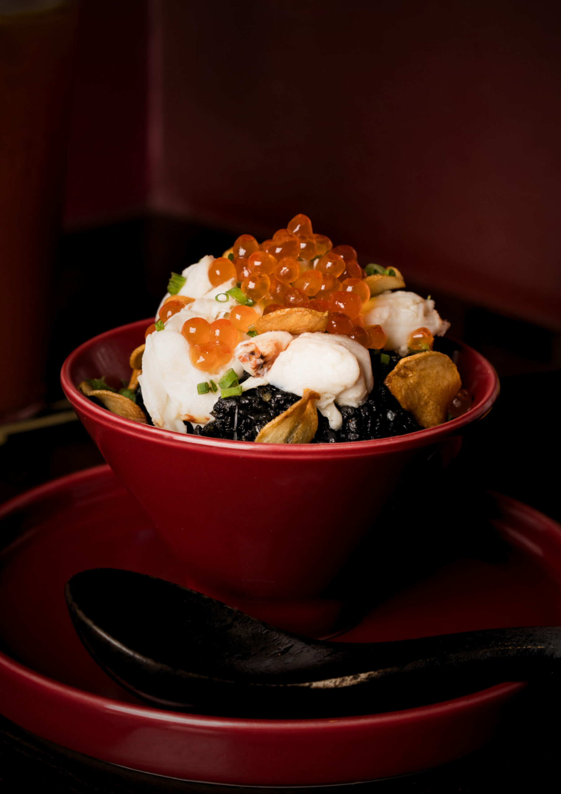 Red bowl of sushi donburi topped with orange salmon roe, creamy white toppings, green onions, and crispy chips against a dark background.