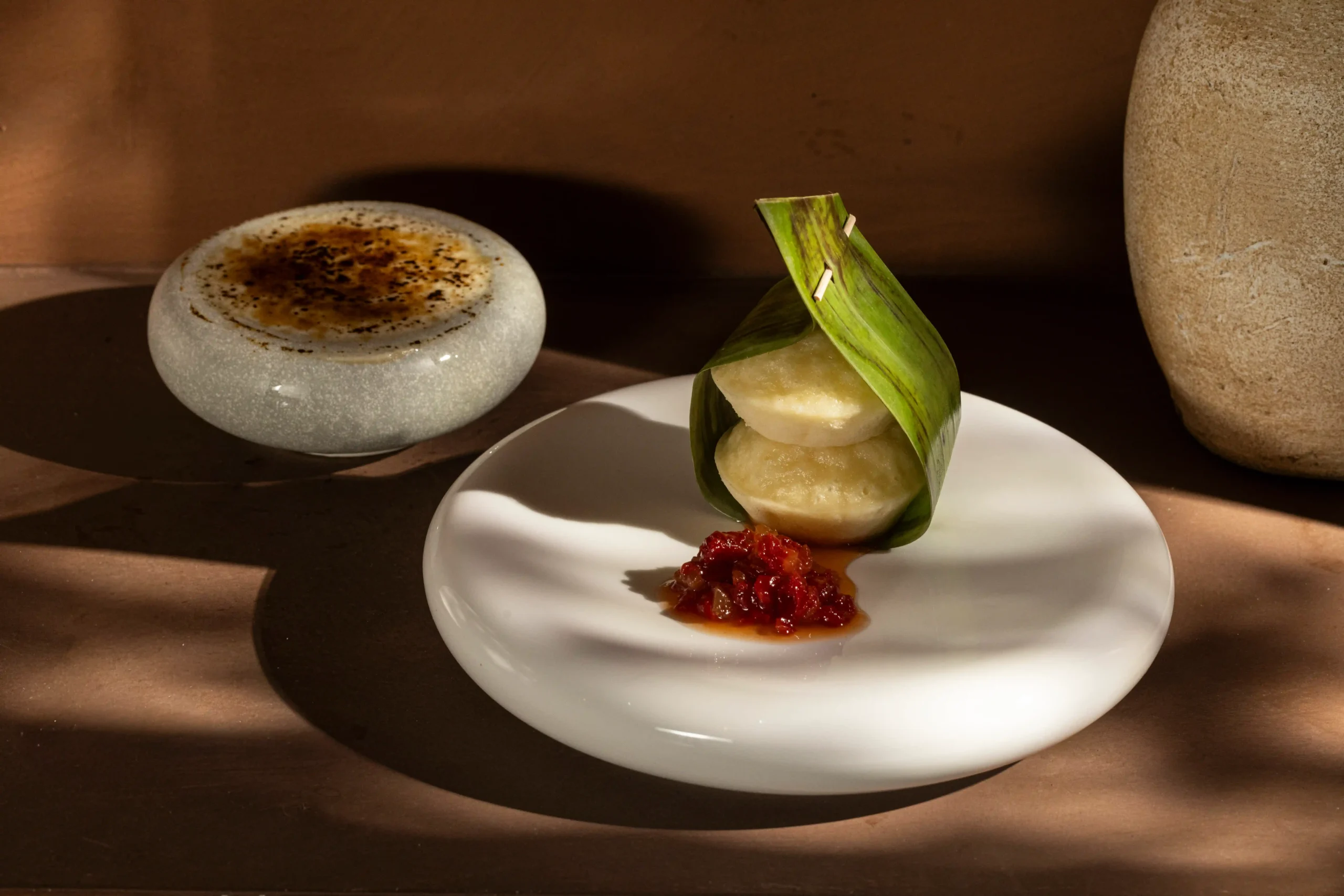 Leaf-wrapped dumpling on a white plate with red relish beside it, set on a wooden table with a stone decor in the background.