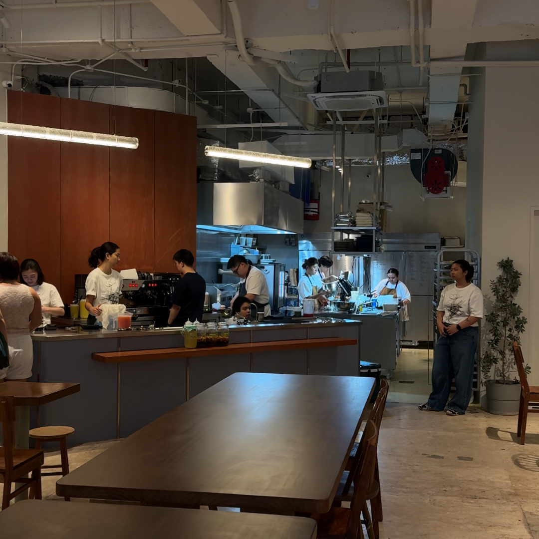 Open kitchen in a modern cafe; staff prep drinks and food behind a long counter, with wooden tables in the foreground.