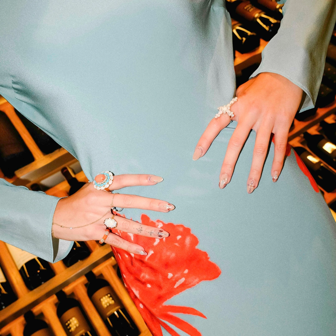 Close-up of a light blue dress with a red floral pattern, showcasing several jeweled rings on both hands and manicured nails.