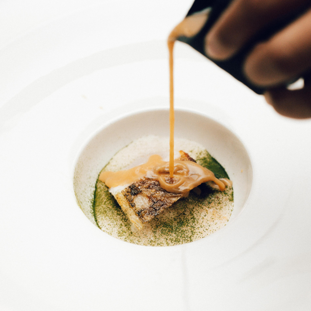 Chef drizzles a smooth brown sauce over a seared fish fillet garnished with green herb powder on a white plate.