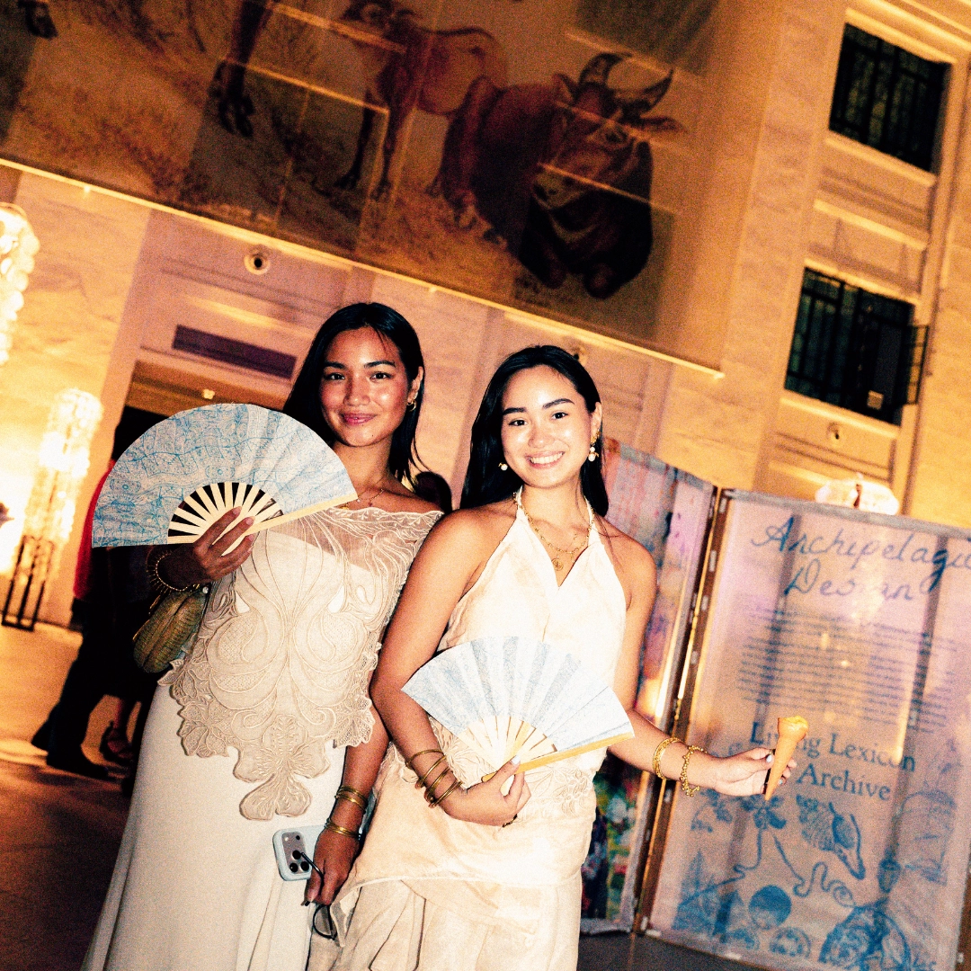 Two young women in cream dresses pose and smile at a night event, each holding a blue hand fan; one also holds a phone in her handly.