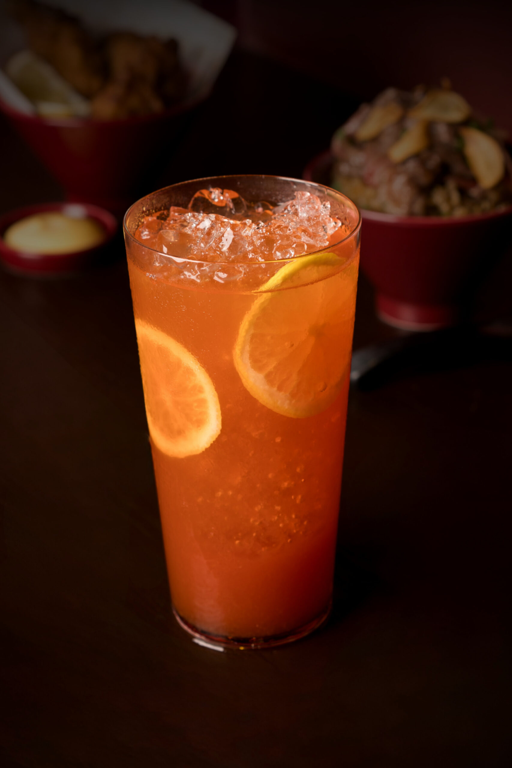 Tall glass of orange cocktail with lemon wedges, crushed ice, and a dark table background. Accurate garnish and setting suggest a refreshing drink.