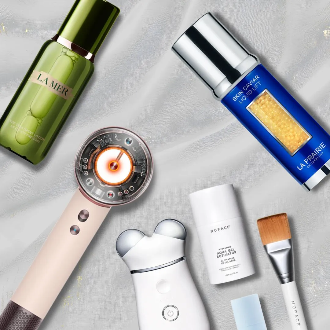 Collection of luxury skincare tools and bottles arranged on a light fabric background, including a green La Mer bottle and a blue La Prairie serum device.