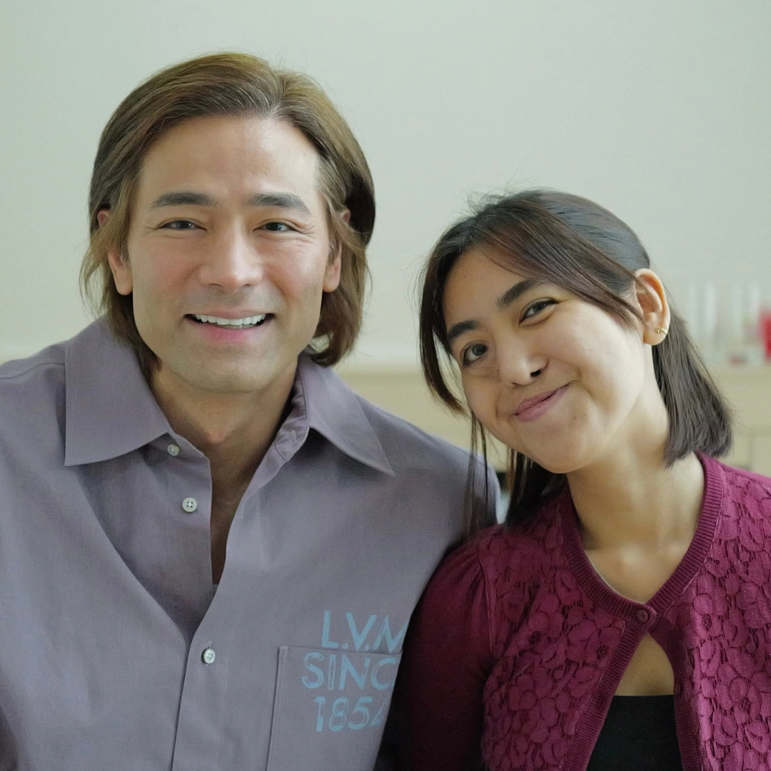 Two people smiling at the camera indoors: man on the left in a lavender shirt and a woman on the right in a burgundy lace cardigan.