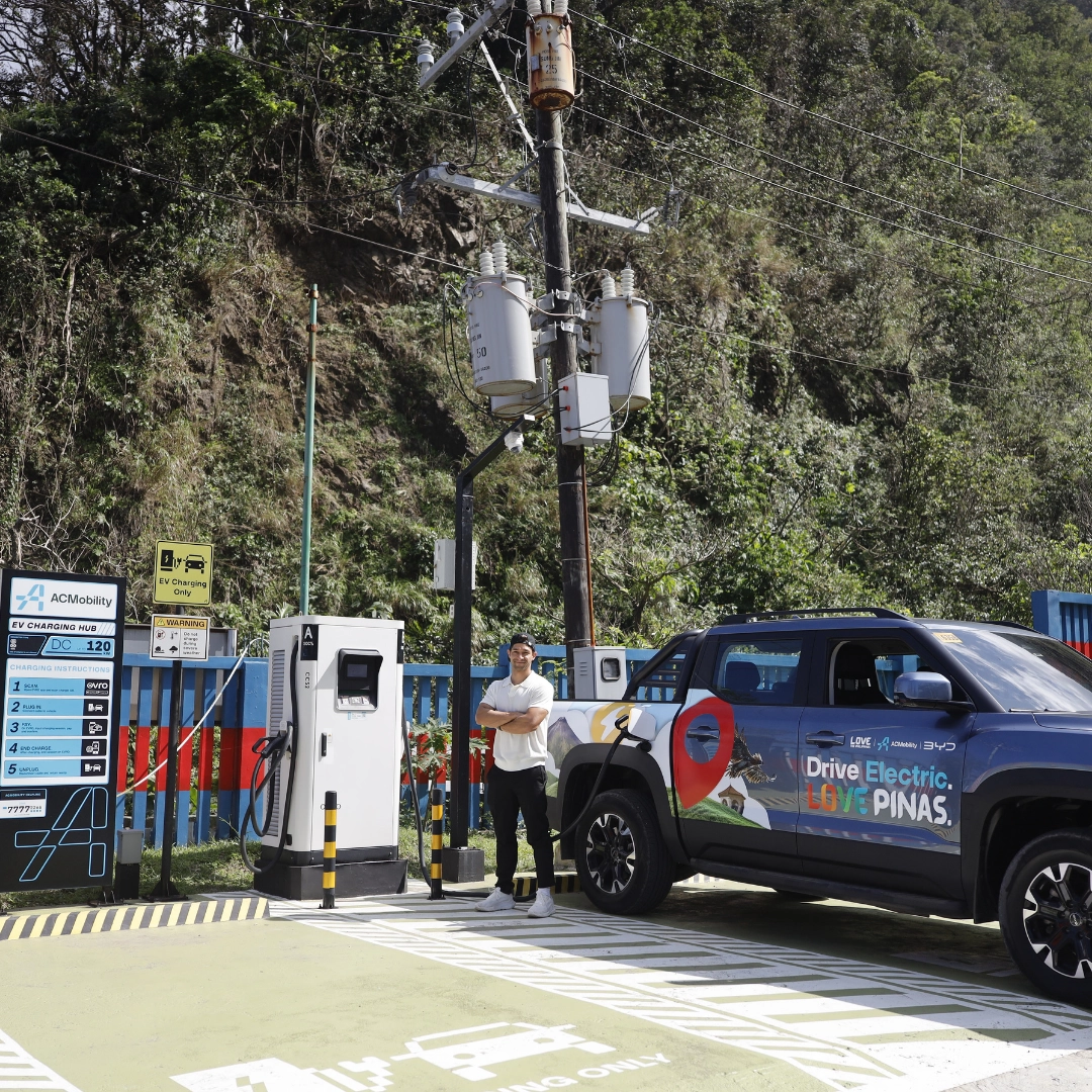 A man stands with arms crossed beside an electric vehicle charging station next to a blue EV with marketing graphics in a charging lot.
